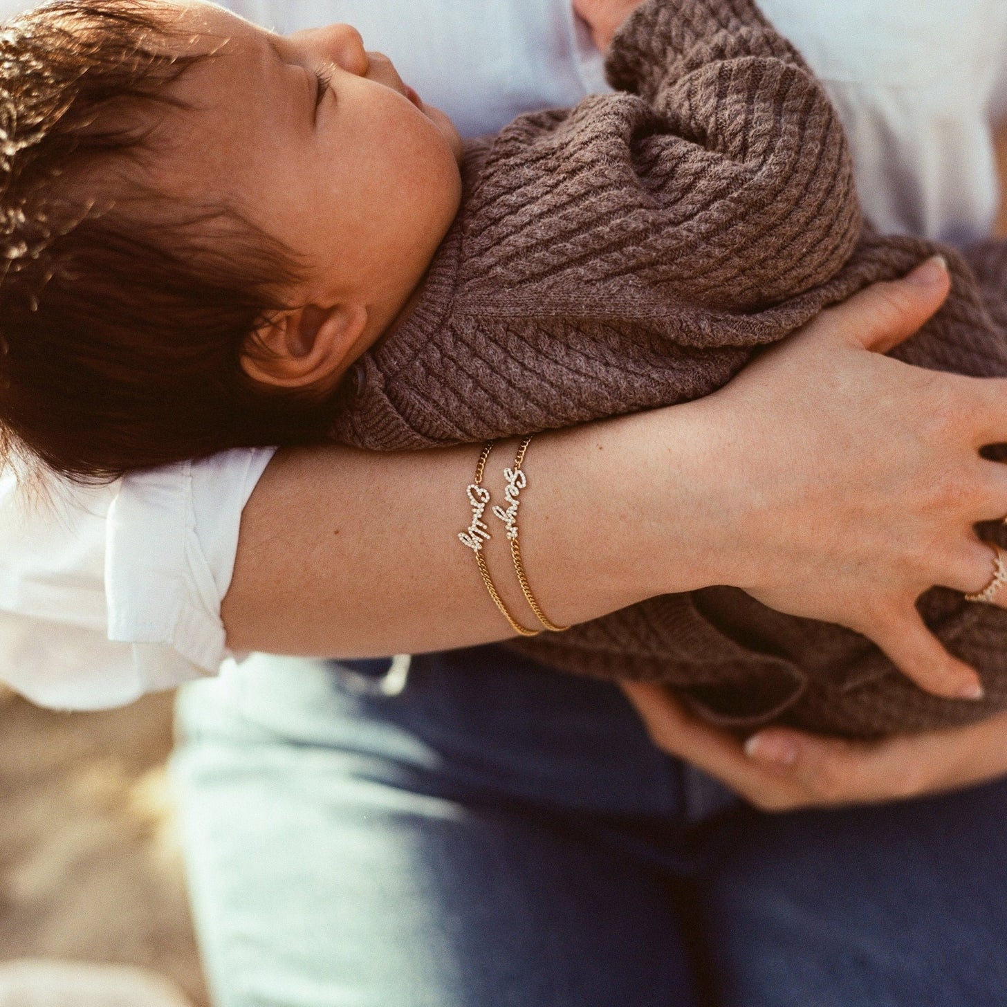Woman holding baby wearing 2 pave name bracelets with curb chains