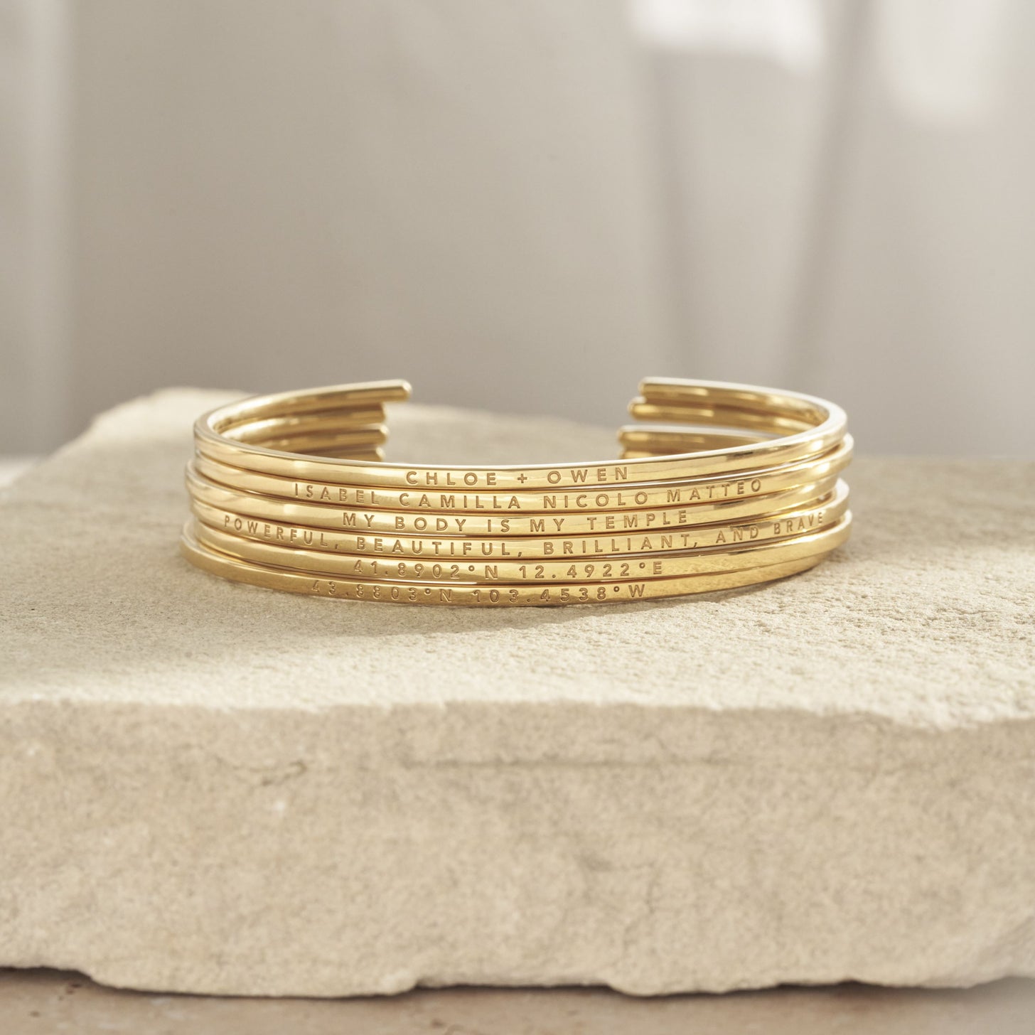 Stacked Rebecca Cuff Bracelets featuring engraved names, messages, and coordinates