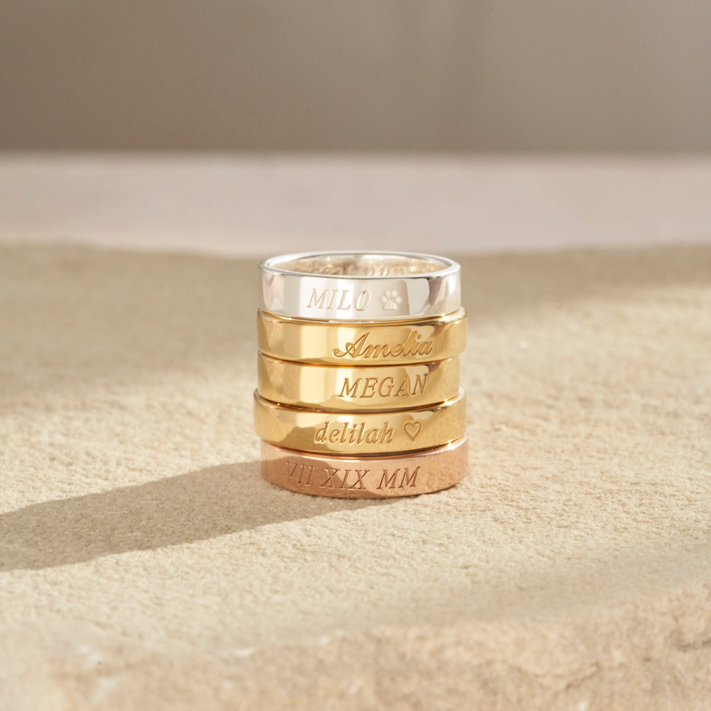 Custom engraved band rings in silver, gold, and rose gold are stacked, showing the elegant italic and cursive fonts
